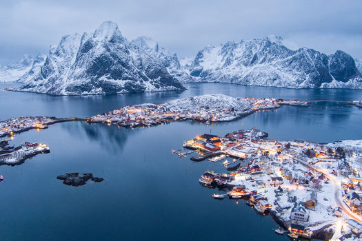 Aerial view of the snowy village of Lofoten with illuminated houses reflecting in the calm water surrounded by dramatic snow covered mountains in Nordland Norway.