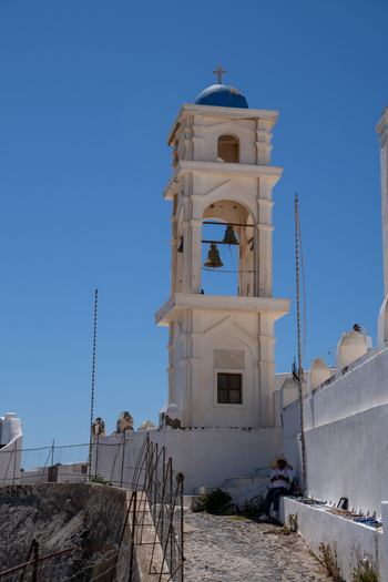 View of a traditional white bell tower with a blue dome and bronze bells against a clear blue sky with a person sitting on a stone path in Fira Greece. View of a traditional white bell tower with a blue dome and bronze bells against a clear blue sky with a person sitting on a stone path in Fira Greece.