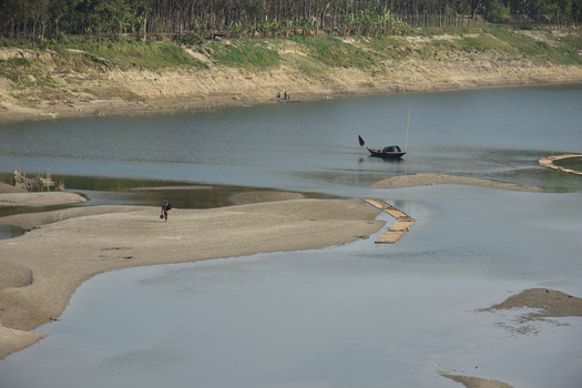 Kushtia District Khulna Division Bangladesh 10 March 2020 View of a person walking on a sandy riverbank with a small boat on the water and trees along the distant shore. Kushtia District Khulna Division Bangladesh 10 March 2020 View of a person walking on a sandy riverbank with a small boat on the water and trees along the distant shore.