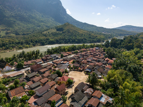 Aerial view of a village with traditional houses lush greenery and a river flowing past mountains in Ban Hat Kho Luang Prabang Province Laos.