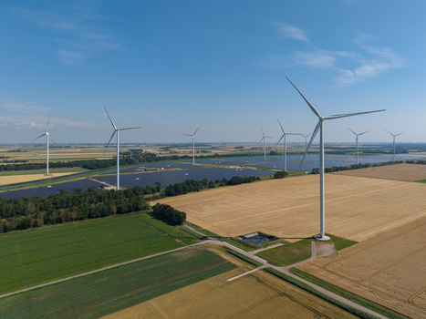 Aerial view of wind turbines and solar panels integrated into a rural landscape with green fields and golden wheat crops under a clear blue sky in the Netherlands.