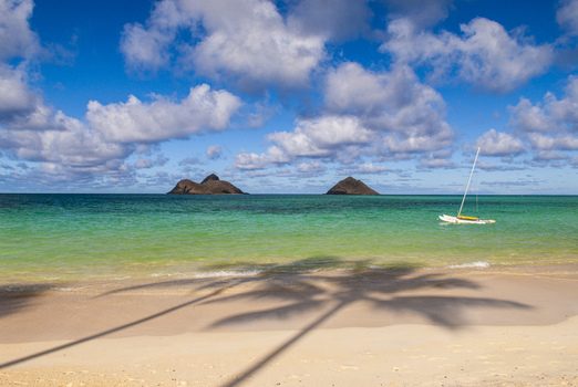View of Lanikai Beach with turquoise water a small sailboat and the Mokolua Islands in the distance under a blue sky with clouds in Kailua Hawaii United States.