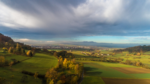 Aerial view of rolling green fields and autumn colored trees surrounding a small town under a dramatic cloudy sky in Germany. Aerial view of rolling green fields and autumn colored trees surrounding a small town under a dramatic cloudy sky in Germany.