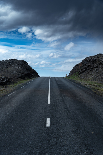 View of an empty asphalt road leading through the rugged volcanic landscape of Hvalfjordur under a dramatic sky with dark storm clouds in Iceland. View of an empty asphalt road leading through the rugged volcanic landscape of Hvalfjordur under a dramatic sky with dark storm clouds in Iceland.