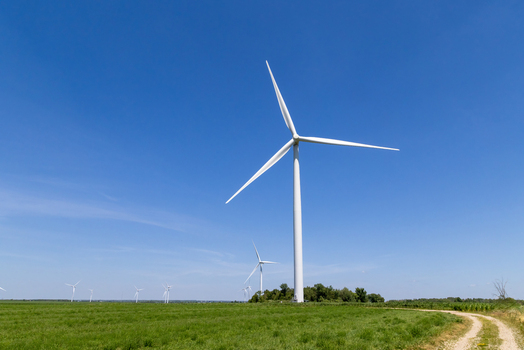 View of large white wind turbines standing in a lush green field under a clear blue sky with a dirt path on the side Toronto Ontario Canada.