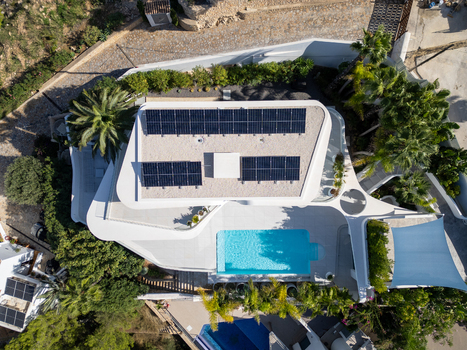 Aerial view of a modern villa with solar panels on the roof and a turquoise swimming pool surrounded by lush palm trees and landscaping in Moraira Valencian Community Spain.