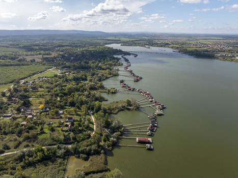 Aerial view of the Bokodi Floating Village with its iconic wooden houses on stilts stretching along the lake shore surrounded by lush greenery and a clear blue sky in Oroszlany Hungary. Aerial view of the Bokodi Floating Village with its iconic wooden houses on stilts stretching along the lake shore surrounded by lush greenery and a clear blue sky in Oroszlany Hungary.