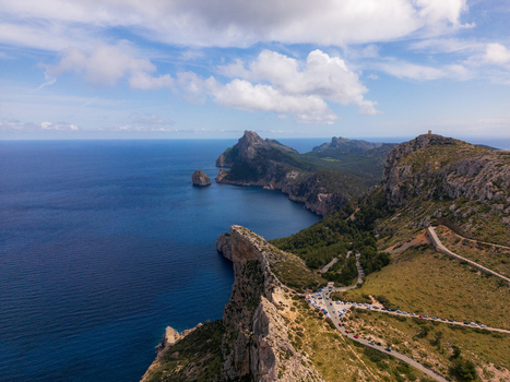 Aerial view of Cap de Formentor featuring rugged limestone cliffs the deep blue Mediterranean Sea and a winding coastal road with parked cars in Balearic Islands Spain.