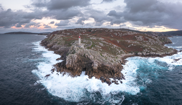 Aerial view of Faro de Punta Nariga on rocky cliffs with crashing white waves and wind turbines under a cloudy sky in Malpica A Coruna Spain. Aerial view of Faro de Punta Nariga on rocky cliffs with crashing white waves and wind turbines under a cloudy sky in Malpica A Coruna Spain.