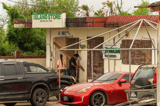 San Jose Tinian Northern Mariana Islands 05 July 2026 View of people working outside a local store with boarded up windows parked cars and tropical foliage. San Jose Tinian Northern Mariana Islands 05 July 2026 View of people working outside a local store with boarded up windows parked cars and tropical foliage.