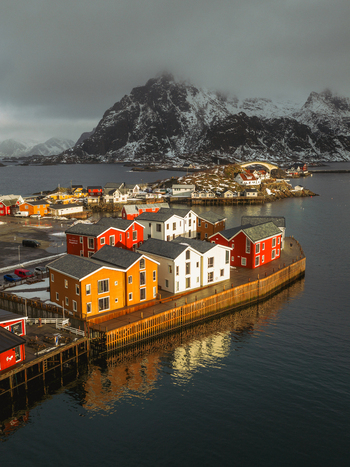 Aerial view of vibrant red orange and white buildings contrast against the moody snow dusted mountain backdrop and dark waters of Henningsv&aelig;r Nordland Norway.