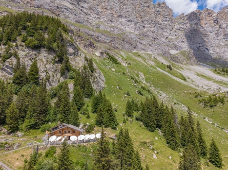 Aerial view of Tallihutte SAC mountain hut nestled on a grassy alpine slope with surrounding pine trees rocky cliffs and white umbrellas on the terrace in Innertkirchen Bern Switzerland.