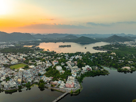 Aerial view of the city of Udaipur situated by Lake Pichola with historic buildings lush greenery and surrounding hills under a warm sunset sky in Udaipur Rajasthan India.
