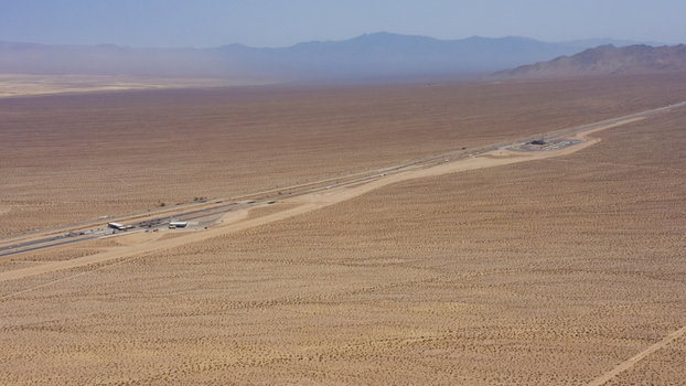 Aerial view of a long highway stretching through the vast arid desert landscape with distant mountains under a clear sky in the United States.