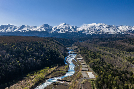 Aerial view of a winding river flowing through a valley with snow capped mountains in the background surrounded by lush forests and small dams in Furano Hokkaido Japan. Aerial view of a winding river flowing through a valley with snow capped mountains in the background surrounded by lush forests and small dams in Furano Hokkaido Japan.