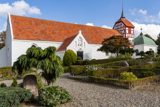 View of the historic church in Hindsholm with white facade red tiled roof and timber framed tower set amidst manicured hedges and a gravel path in Kerteminde Municipality Denmark. View of the historic church in Hindsholm with white facade red tiled roof and timber framed tower set amidst manicured hedges and a gravel path in Kerteminde Municipality Denmark.