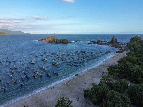 Aerial view of Papuma Beach with numerous traditional fishing boats anchored in the bay sandy coastline lush green trees and rocky outcrops in Jember East Java Indonesia. Aerial view of Papuma Beach with numerous traditional fishing boats anchored in the bay sandy coastline lush green trees and rocky outcrops in Jember East Java Indonesia.