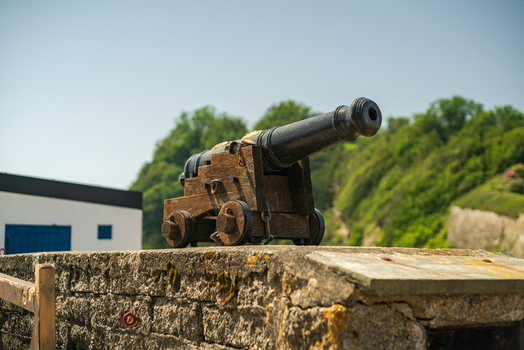 View of a historic iron cannon mounted on a wooden carriage resting on a weathered stone wall in the harbor of Charlestown Cornwall United Kingdom. View of a historic iron cannon mounted on a wooden carriage resting on a weathered stone wall in the harbor of Charlestown Cornwall United Kingdom.