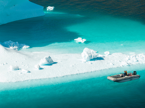 Aerial view of a zodiac boat with people navigating through turquoise water near a large white iceberg in Greenland. Aerial view of a zodiac boat with people navigating through turquoise water near a large white iceberg in Greenland.