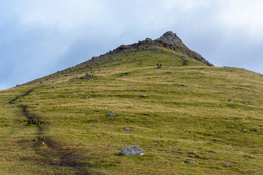View of a grassy mountain slope with a hiking trail leading towards a rocky peak in the Faroe Islands. View of a grassy mountain slope with a hiking trail leading towards a rocky peak in the Faroe Islands.
