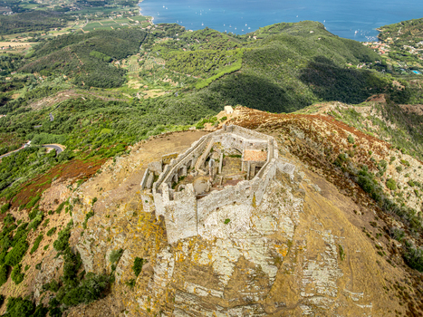 Aerial view of Castello del Volterraio an ancient stone fortress ruin perched on a rocky hilltop surrounded by lush Mediterranean hills and coastal scenery in Portoferraio Toscana Italy.