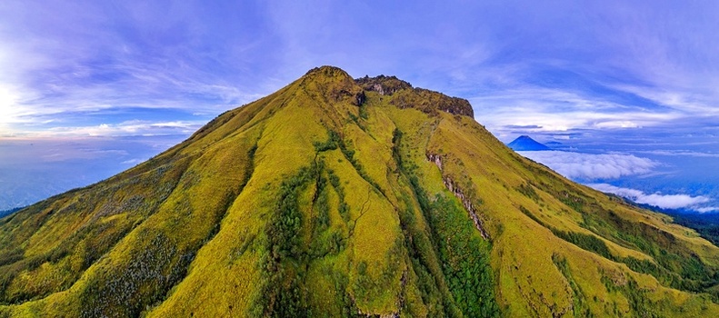 Aerial view of the lush green slopes and volcanic peak of Mount Sumbing under a blue sky with a sea of clouds in Temanggung Central Java Indonesia. Aerial view of the lush green slopes and volcanic peak of Mount Sumbing under a blue sky with a sea of clouds in Temanggung Central Java Indonesia.