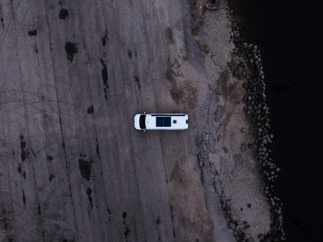 Aerial view of a white camper van parked on a gravel shore next to a dark body of water in Latvia.