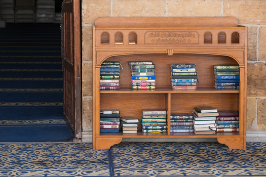View of a wooden bookshelf filled with stacks of religious books on a blue patterned carpet at the Al Azhar Mosque Cairo Egypt. View of a wooden bookshelf filled with stacks of religious books on a blue patterned carpet at the Al Azhar Mosque Cairo Egypt.