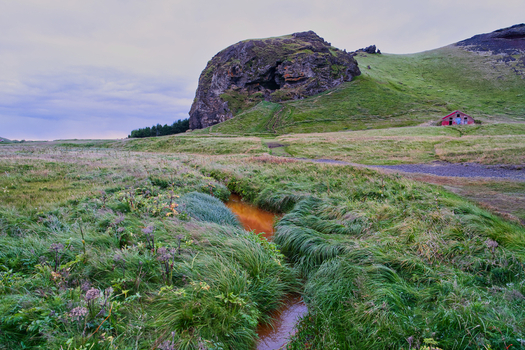 View of Dyrholaey rock formation with a small red building a winding stream with orange water and lush green grassy fields in Vik Myrdalshreppur Iceland. View of Dyrholaey rock formation with a small red building a winding stream with orange water and lush green grassy fields in Vik Myrdalshreppur Iceland.