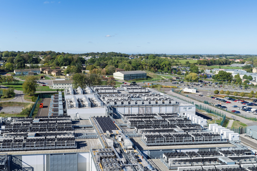 Aerial view of a large data center facility featuring numerous cooling units on the roof surrounded by parking areas and green landscape in the United Kingdom.