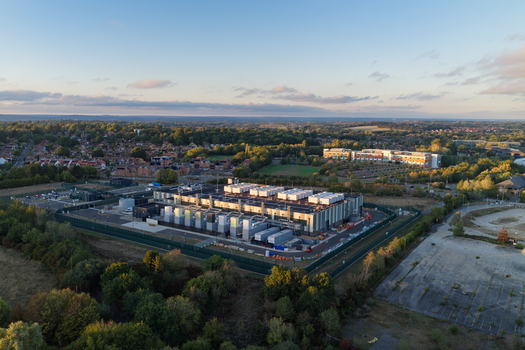 Aerial view of a modern data center facility surrounded by lush green trees and suburban residential areas in the United Kingdom. Aerial view of a modern data center facility surrounded by lush green trees and suburban residential areas in the United Kingdom.