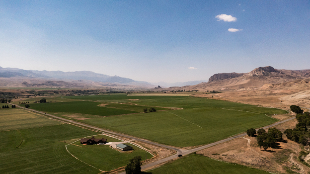 Aerial view of the rural landscape near Cody featuring green agricultural fields a farmstead a road and distant arid mountains under a clear blue sky in Cody Wyoming United States.