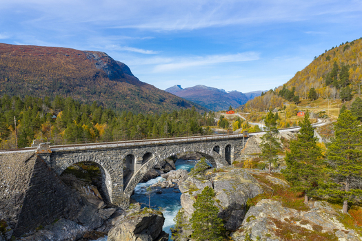Aerial view of a historic stone arch railway bridge spanning a rushing river in a mountainous landscape with autumn foliage and clear blue sky in Bjorli Innlandet Norway. Aerial view of a historic stone arch railway bridge spanning a rushing river in a mountainous landscape with autumn foliage and clear blue sky in Bjorli Innlandet Norway.