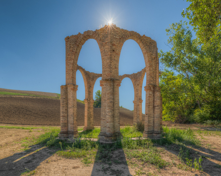View of a historic brick arch structure standing in a rural field under a bright sunburst in Ponzano di Fermo Marche Italy. View of a historic brick arch structure standing in a rural field under a bright sunburst in Ponzano di Fermo Marche Italy.