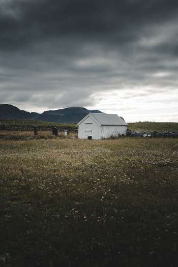 View of a small white wooden building standing in a grassy field under a dramatic cloudy sky in Ramberg Nordland Norway. View of a small white wooden building standing in a grassy field under a dramatic cloudy sky in Ramberg Nordland Norway.