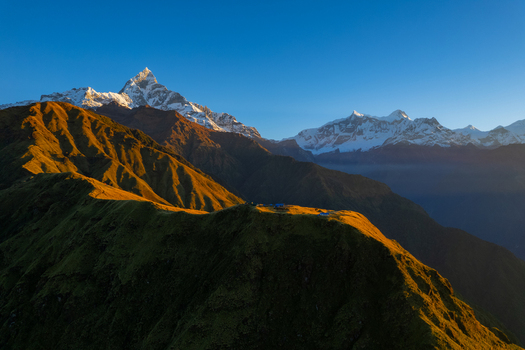 Aerial view of the Himalayan mountain range with snow capped peaks and golden hour light illuminating the grassy ridges in Nepal.