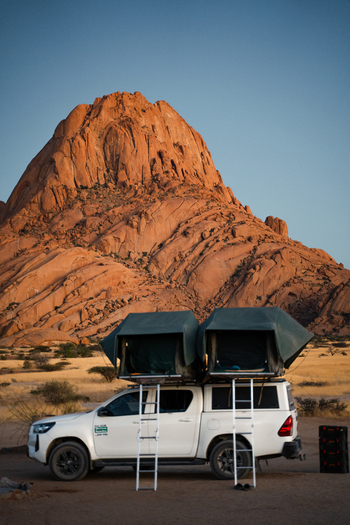 Spitzkoppe Namibia 12 March 2026 View of a white vehicle with a roof tent stands poised against the backdrop of the rugged sun kissed Spitzkoppe mountains.