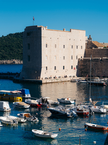View of St. John Fortress and the Old Port with small boats moored in the harbor under a clear blue sky in Dubrovnik Dubrovnik Neretva Croatia. View of St. John Fortress and the Old Port with small boats moored in the harbor under a clear blue sky in Dubrovnik Dubrovnik Neretva Croatia.