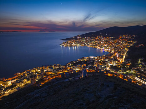 Aerial view of the coastal city of Saranda illuminated at night with city lights reflecting on the Ionian Sea under a twilight sky in Saranda Vlore County Albania. Aerial view of the coastal city of Saranda illuminated at night with city lights reflecting on the Ionian Sea under a twilight sky in Saranda Vlore County Albania.