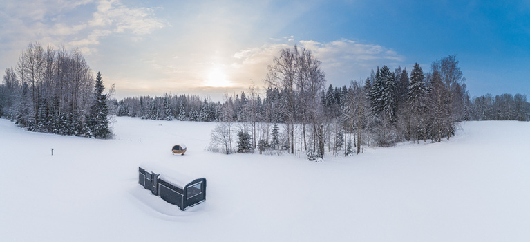 Aerial view of a modern cabin and a barrel sauna in a snow covered field surrounded by a forest under a bright winter sky in Latvia. Aerial view of a modern cabin and a barrel sauna in a snow covered field surrounded by a forest under a bright winter sky in Latvia.