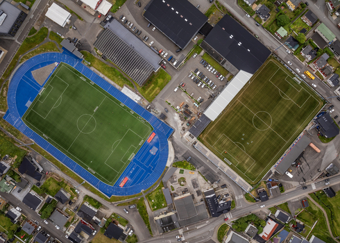 Aerial view of Djupumyra Stadium featuring green football pitches and a blue running track surrounded by urban houses and parking lots in Klaksvik Northern Isles Faroe Islands.