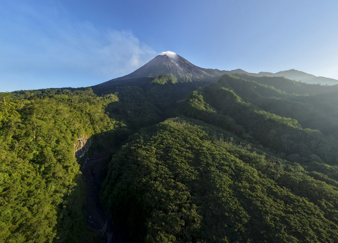 Aerial view of Mount Merapi volcano with a smoke plume at the summit and lush green forest slopes under a clear blue sky in Kecamatan Pakem Daerah Istimewa Yogya Indonesia. Aerial view of Mount Merapi volcano with a smoke plume at the summit and lush green forest slopes under a clear blue sky in Kecamatan Pakem Daerah Istimewa Yogya Indonesia.