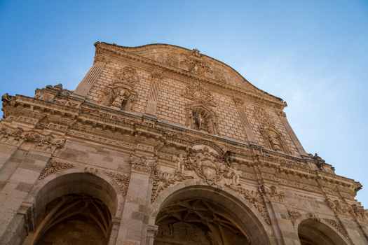 View of the ornate stone facade of a historic church with intricate carvings and arched entrances under a clear blue sky in Sardinia Italy.