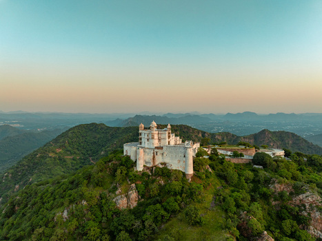 Aerial view of Sajjangarh Palace perched on a hilltop surrounded by lush green forests and rolling hills at sunset in Udaipur Rajasthan India.