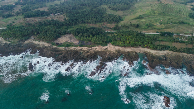 Aerial view of a rocky coastline with crashing waves lush tropical vegetation and terraced fields in Indonesia. Aerial view of a rocky coastline with crashing waves lush tropical vegetation and terraced fields in Indonesia.