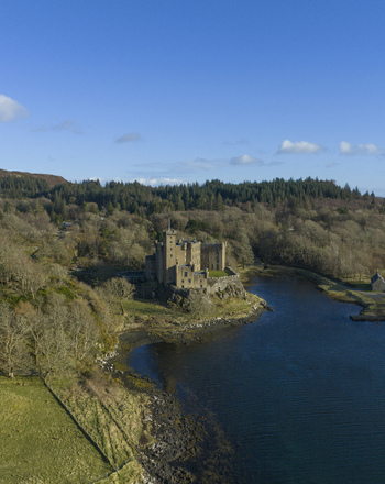 Aerial view of Dunvegan Castle and Gardens on a rocky cliff overlooking the water surrounded by dense woodland and rolling green hills in Isle of Skye Scotland United Kingdom. Aerial view of Dunvegan Castle and Gardens on a rocky cliff overlooking the water surrounded by dense woodland and rolling green hills in Isle of Skye Scotland United Kingdom.