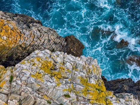 Aerial view of Silence Beach Viewpoint with rugged cliffs covered in yellow lichen meeting the crashing turquoise waves in Cudillero Asturias Spain.