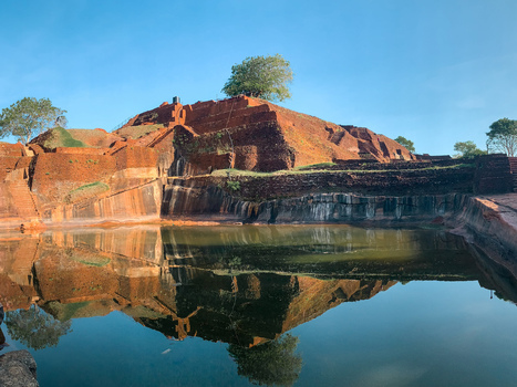 View of the ancient Sigiriya rock fortress reflected in the calm water of the royal gardens with red brick ruins and a clear blue sky Sri Lanka.