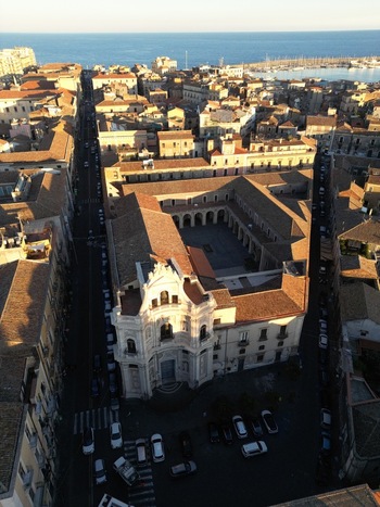 Aerial view of the historic Church of San Placido and its courtyard surrounded by traditional rooftops and a long street leading to the harbor under golden hour light in Catania Sicily Italy. Aerial view of the historic Church of San Placido and its courtyard surrounded by traditional rooftops and a long street leading to the harbor under golden hour light in Catania Sicily Italy.