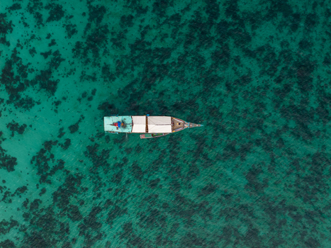 Aerial view of a traditional wooden phinisi boat in the turquoise waters of Komodo National Park with coral reefs visible beneath the surface in Komodo East Nusa Tenggara Indonesia. Aerial view of a traditional wooden phinisi boat in the turquoise waters of Komodo National Park with coral reefs visible beneath the surface in Komodo East Nusa Tenggara Indonesia.
